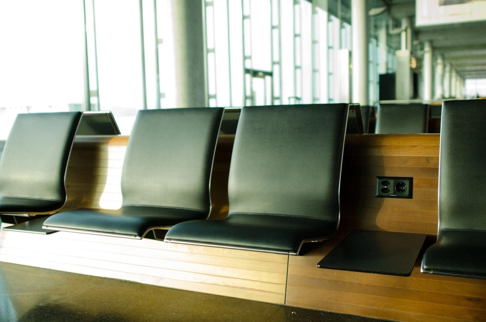 Four modern black chairs with wooden backing in an empty area next to a power outlet, situated in a sunlit room with large windows.