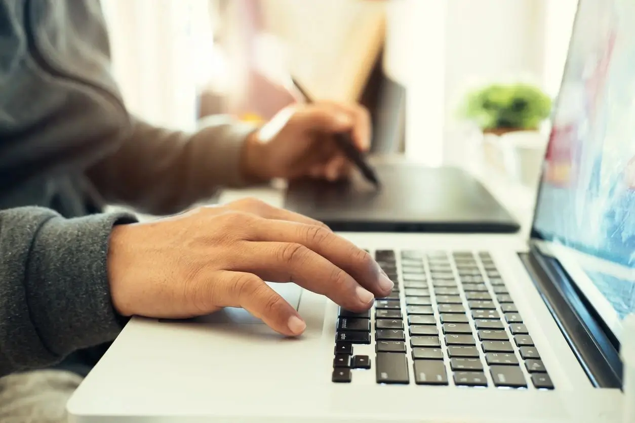 Person typing on a laptop with a tablet and stylus nearby.