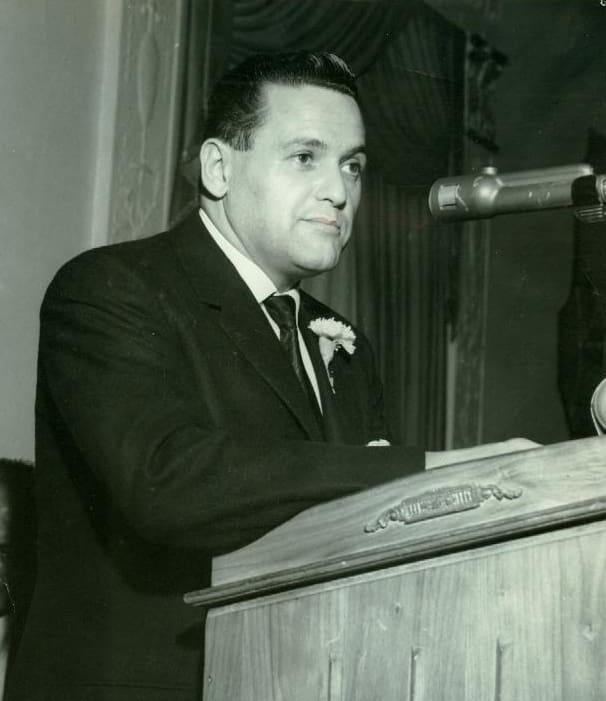 A man in a suit speaking at a podium with a microphone.