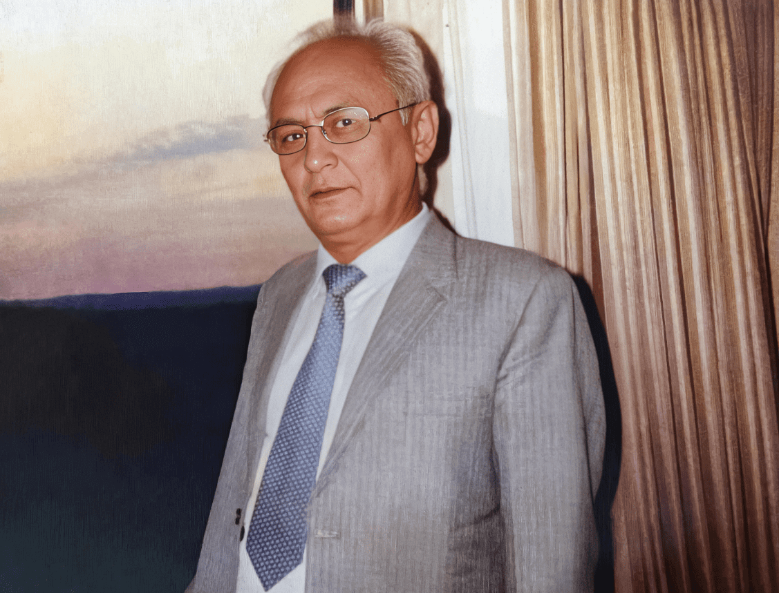 Elderly man in suit near window curtains.